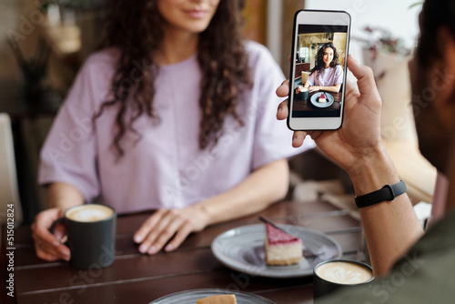 Hand of guy with smartphone taking photo of his girlfriend with cup of coffee and cake sitting by table in cafe or restaurant photo