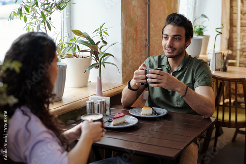 Guy with cup of coffee and his girlfriend sitting by table in front of one another and chatting while having coffee with tasty dessert