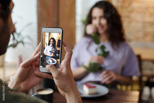 Hand of guy with smartphone taking photo of his girlfriend with rose and cheesecake sitting by table in front of him during romantic date