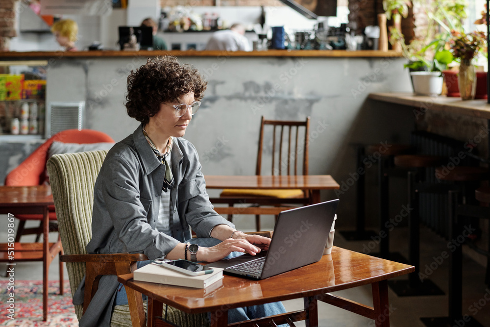 Young serious businesswoman in casualwear concentrating on organization of work while typing on laptop keyboard in cafe Young serious businesswoman in casualwear concentrating on organization of work while typing on laptop keyboard in cafe