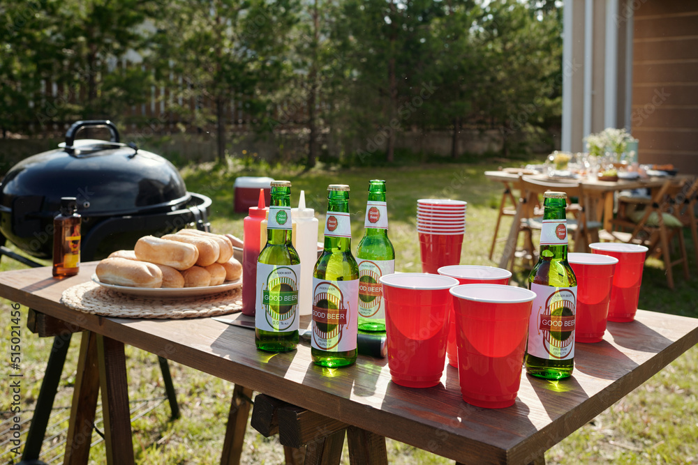 Obraz premium Wooden table with homemade food, bottles of beer, plastic cups, ketchup prepared for barbeque party standing in front of country house