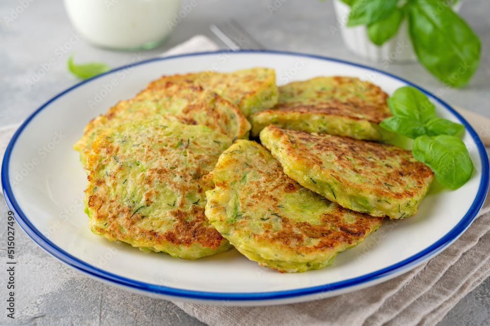 Zucchini fritters with cheese, garlic and herbs. Vegetarian zucchini pancakes, served with basil and sour cream on a gray concrete background. Selective focus. Zucchini fritters with cheese, garlic and herbs. Vegetarian zucchini pancakes, served with basil and sour cream on a gray concrete background. Selective focus.