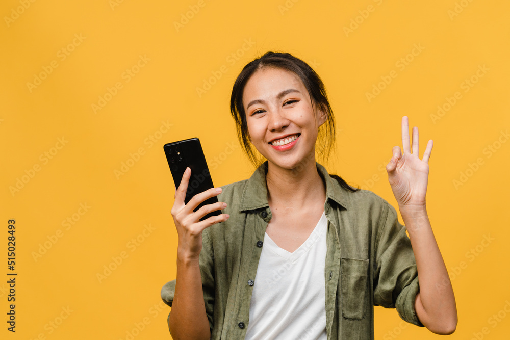 Surprised young Asia lady using mobile phone with positive expression, smile broadly, dressed in casual clothing and looking at camera on yellow background. Happy adorable glad woman rejoices success. Surprised young Asia lady using mobile phone with positive expression, smile broadly, dressed in casual clothing and looking at camera on yellow background. Happy adorable glad woman rejoices success.