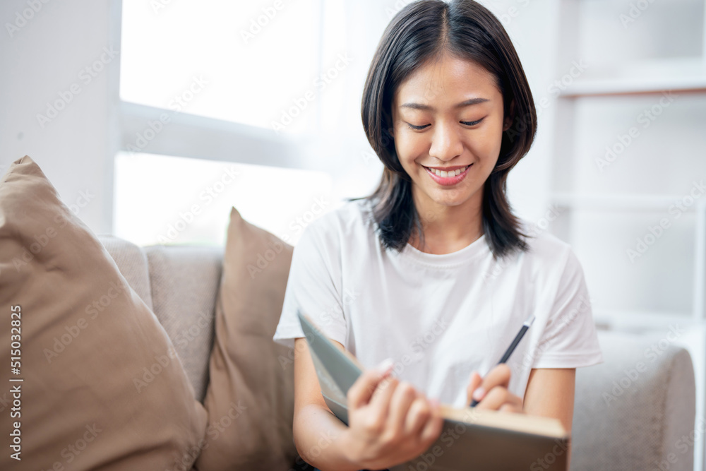 Young asian woman writing on notebook sitting on couch at home.. Young asian woman writing on notebook sitting on couch at home..
