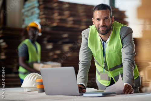 Happy businessman working on laptop at wood warehouse and looking at camera. © Drazen