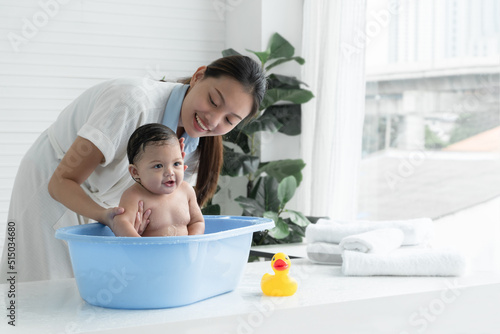 Fotografie, Obraz Happy Asian little baby sitting and enjoy playing water in bathtub while young mother wear bathrobe is bathing her cute daughter at home