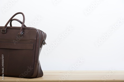 An elegant business briefcase made of old craft dark brown leather stands next to the white wall as a background on the smooth surface of light wooden table. No people studio photography.