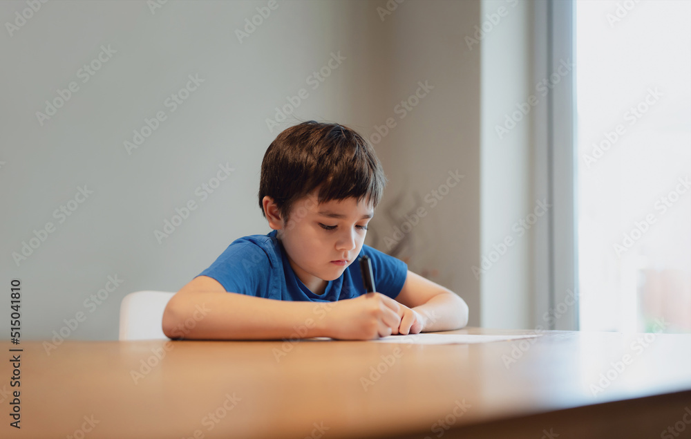Kid siting on table doing homework,Child boy holding black pen writing on white paper,Young boy practicing English words at home. Elementary school and home schooling, Distance Education concept Kid siting on table doing homework,Child boy holding black pen writing on white paper,Young boy practicing English words at home. Elementary school and home schooling, Distance Education concept