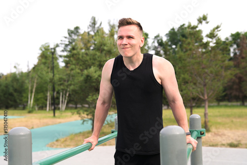 Young handsome fit athletic strong man is training doing triceps and chest pull ups in park on sports ground outdoors on bar at summer. Open air gym. Bodybuilding, fitness sport healthy lifestyle.