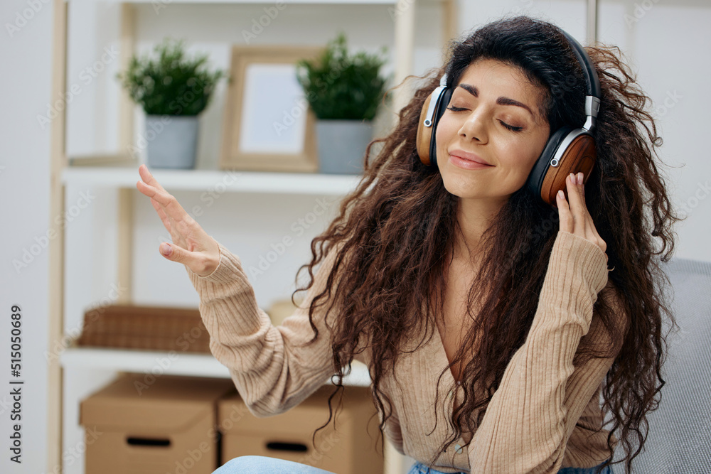 Relaxed calm pleasant tanned curly Latin lady with headphones moves to cool music sit on chair in home modern interior close eyes. Copy space Mockup Banner. Concept meditation. Relax in home Relaxed calm pleasant tanned curly Latin lady with headphones moves to cool music sit on chair in home modern interior close eyes. Copy space Mockup Banner. Concept meditation. Relax in home