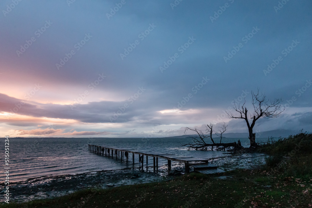 Long exposure view of a pier on a lake at dusk, with perfectly still water, moving clouds and skeletal trees