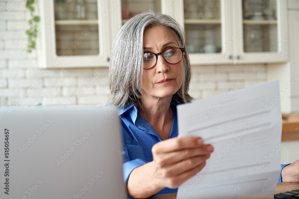 Serious worried mature woman reading paper bill pay online at home. Old lady holding bank letter managing account finances, calculating taxes, planning loan debt pension payment sit at kitchen table. - obrazy, fototapety, plakaty 