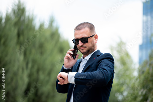 A business man in dark sunglasses with a phone in his hands stands outside the business building talks on the phone and looks at the watch. He is dressed in a light shirt and casual style jacket.