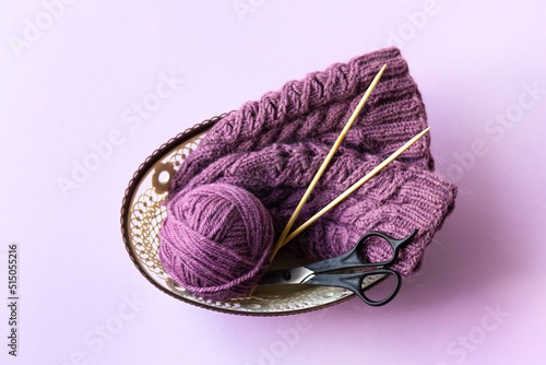 A purple ball of thread and a knitted hat with knitting needles and scissors in a basket on a purple background, knitting hobby photo