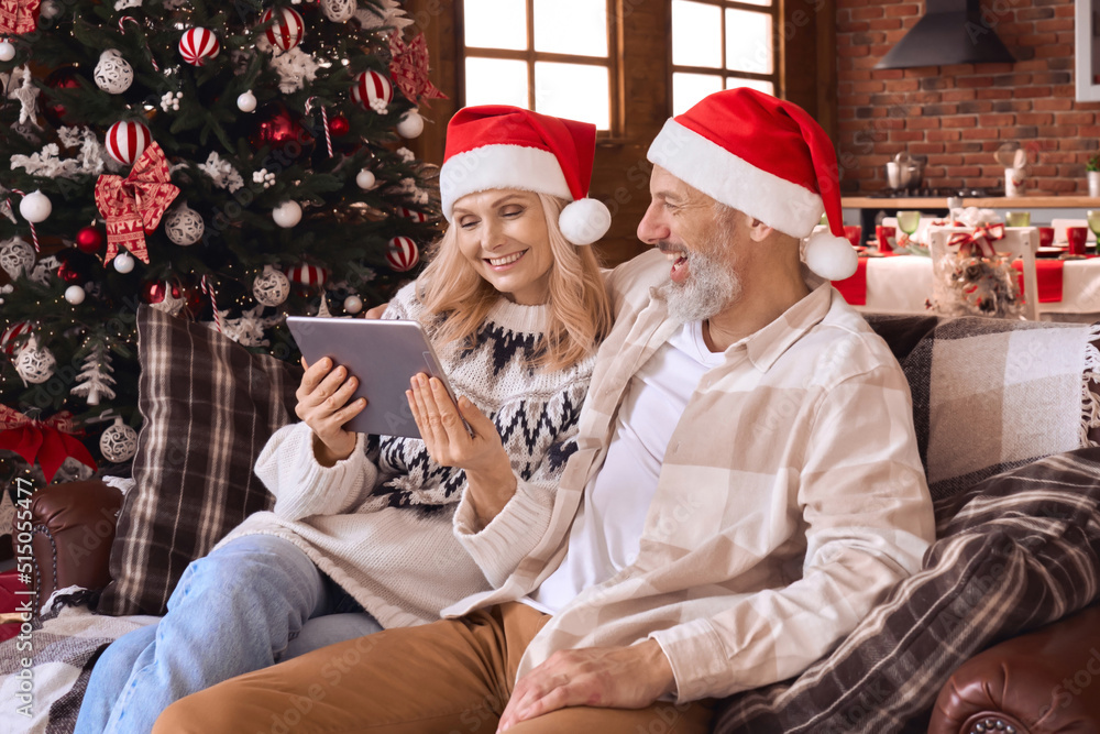 Happy 50s older couple using digital tablet in living room on Christmas. Smiling mature grandparents wearing santa hats doing holiday online shopping having family video call relaxing on sofa at home.