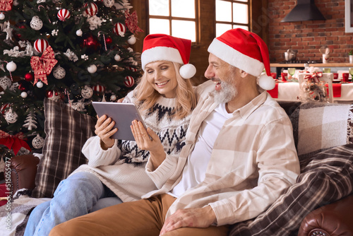 Happy 50s older couple using digital tablet in living room on Christmas. Smiling mature grandparents wearing santa hats doing holiday online shopping having family video call relaxing on sofa at home. © insta_photos