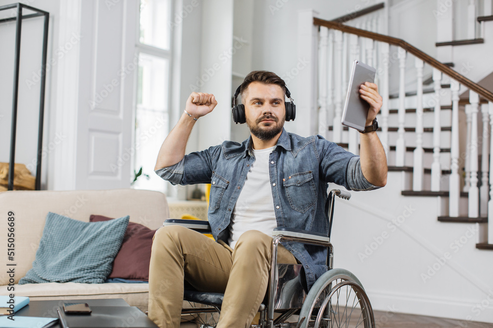 Handsome wheelchair user in headphones dancing with hands to nice music in modern apartment. Bearded man with disability holding digital tablet for choosing new track. Handsome wheelchair user in headphones dancing with hands to nice music in modern apartment. Bearded man with disability holding digital tablet for choosing new track.
