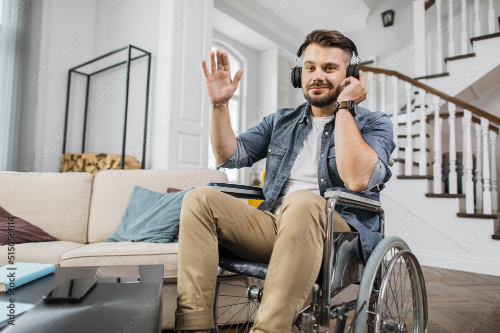 Disabled man in modern headphones waving hand while looking at camera with smile. Pleasant guy sitting in wheelchair and listening good music at home.
