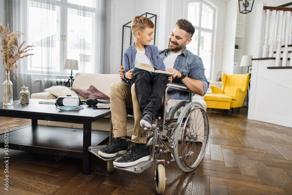 School age child sitting on fathers knees in wheelchair and preparing for school lesson while reading educational book at home. Disabled dad helping concentrated son finishing homework.