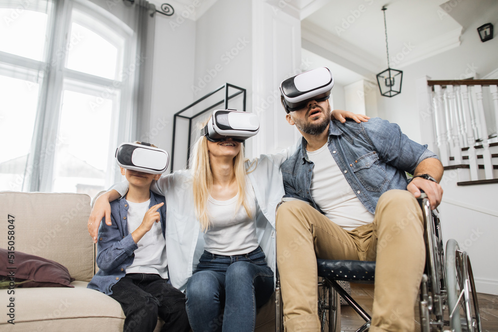 Attractive mother hugging cute son and bearded father in wheelchair while using vr headset at cozy home. Concept of happy family, modern technology and leisure time. Attractive mother hugging cute son and bearded father in wheelchair while using vr headset at cozy home. Concept of happy family, modern technology and leisure time.