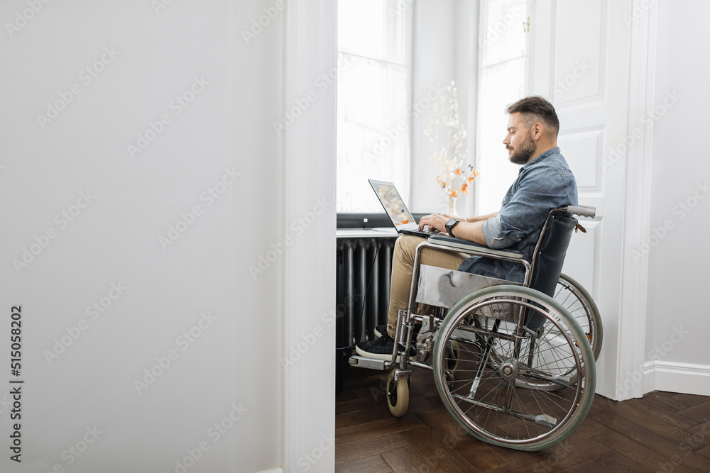 Male freelancer with disability working on wireless laptop in bright room at home. Bearded man wearing casual clothes sitting in wheelchair near big window. Male freelancer with disability working on wireless laptop in bright room at home. Bearded man wearing casual clothes sitting in wheelchair near big window.