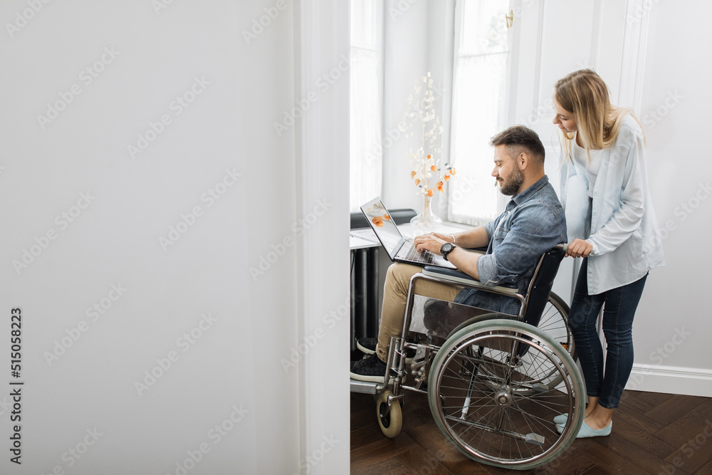 Disabled caucasian man sitting in wheelchair and working on modern laptop while his charming wife standing behind. Family support, people and love. Disabled caucasian man sitting in wheelchair and working on modern laptop while his charming wife standing behind. Family support, people and love.