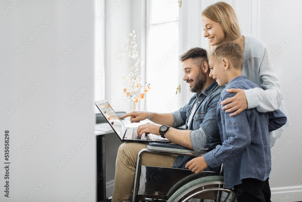 Happy mother embracing smiling son and satisfied bearded husband which sitting in wheelchair at home. Friendly loving family using laptop together near window.