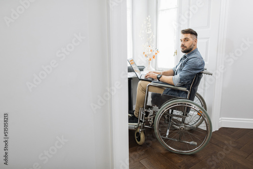 Focused bearded man who using wheelchair holding laptop on knees and looking at camera. Hardworking male freelancer with disability working remotely from home.