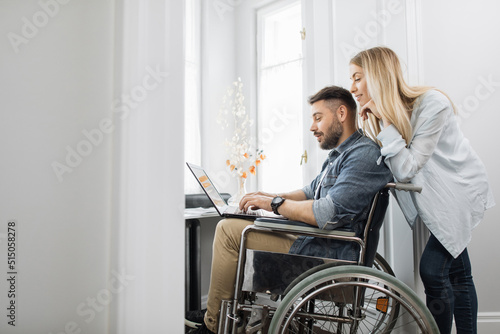Bearded young man in wheelchair looking at laptop screen while beautiful wife standing behind at home office. Happy wife supporting beloved disabled husband.