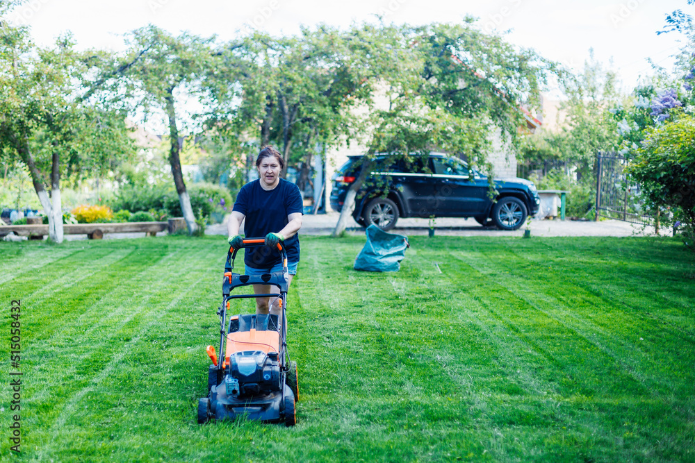 Dark-haired female gardener in casual clothes using lawn mower or grass cutter in the yard, grounds. Technological machine for lawns. Cutting fresh lush green grass in countryside. Grass equipment Dark-haired female gardener in casual clothes using lawn mower or grass cutter in the yard, grounds. Technological machine for lawns. Cutting fresh lush green grass in countryside. Grass equipment