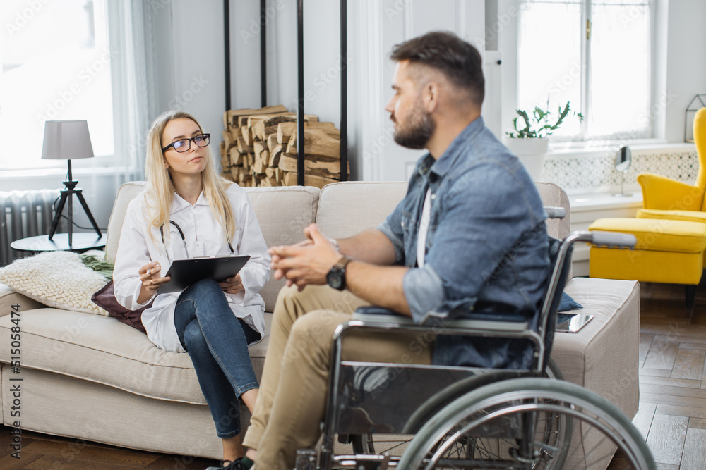 Caring female doctor with medical documents consulting disabled man at living room. Young male patient in wheelchair listening prescription from therapist.