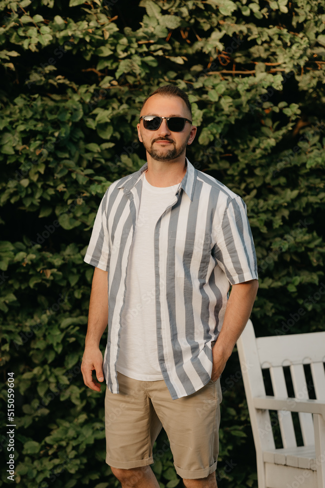 A smiling man is posing in the garden in the evening. A happy guy in sunglasses with green leaves in the background A smiling man is posing in the garden in the evening. A happy guy in sunglasses with green leaves in the background