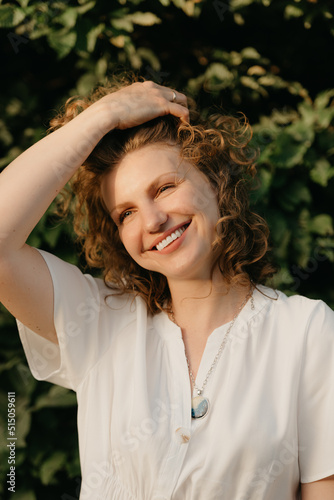A smiling woman with curly hair is posing in the park in the evening. A lady in a white dress with her arm on her head with green leaves in the background.