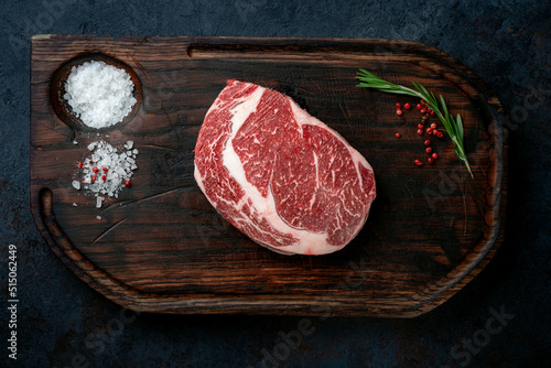 Raw rib eye steak of marbled beef on a wooden board with spices and herbs for grilling.