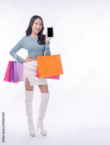 Cheerful excited asian woman showing mobile phone screen carrying shopping bags full body standing on white background. Joyful model young girl surprise application shopping online over isolated.