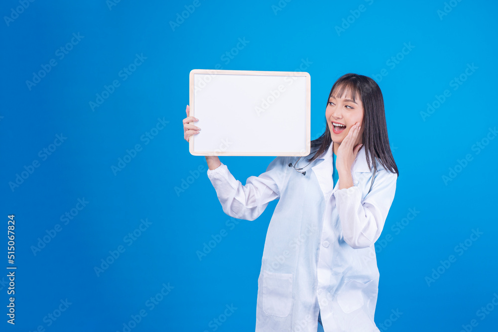 Friendly Asian doctor young woman wear uniform coat with stethoscope presenting blank white board while standing over isolated blue background. Physician health care or insurance concept.. Friendly Asian doctor young woman wear uniform coat with stethoscope presenting blank white board while standing over isolated blue background. Physician health care or insurance concept..