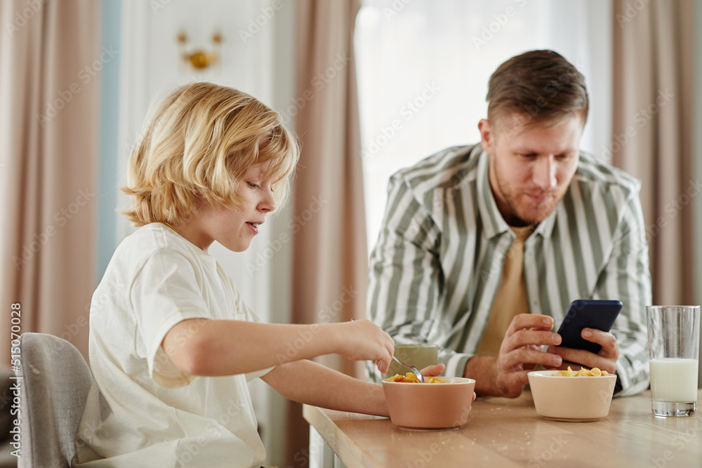 Side view portrait of father and son at breakfast in morning with dad using smartphone in background, copy space Side view portrait of father and son at breakfast in morning with dad using smartphone in background, copy space