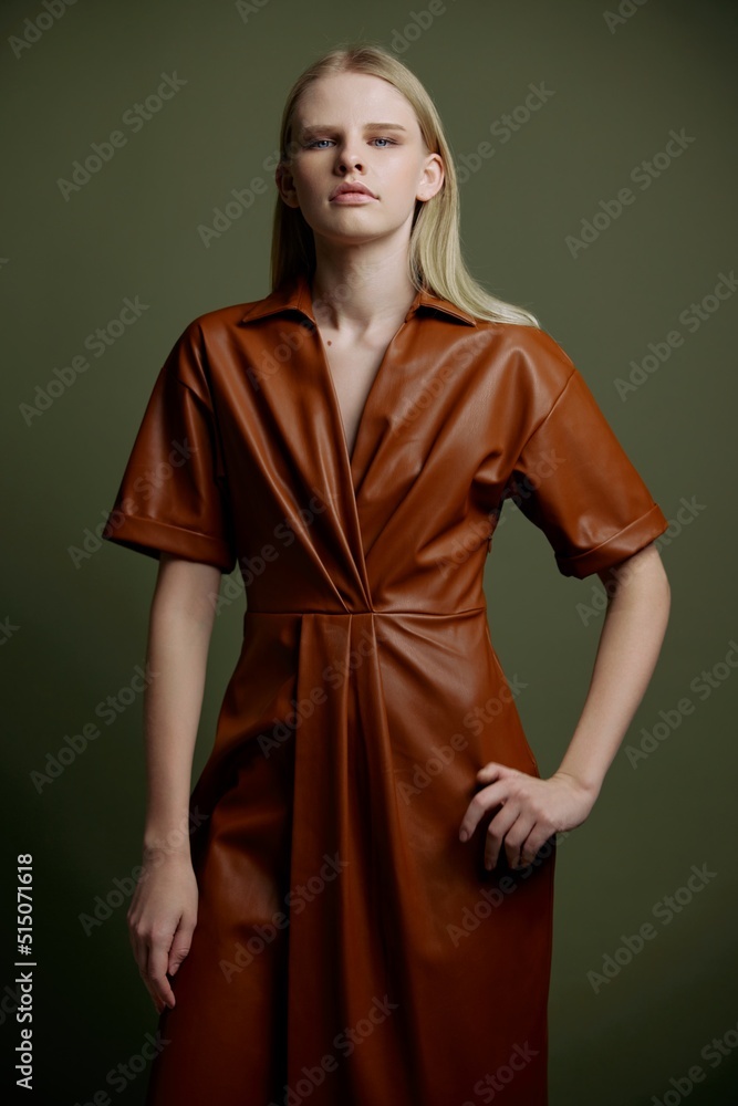 Trendy stylish offer for your brand. Fashion gorgeous young blonde lady holding hand on waist dress in leather trench coat serious looks at camera posing isolated on over olive green studio background Trendy stylish offer for your brand. Fashion gorgeous young blonde lady holding hand on waist dress in leather trench coat serious looks at camera posing isolated on over olive green studio background