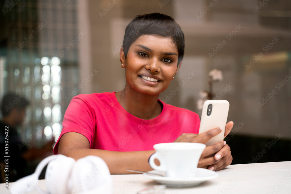 Portrait of smiling Indian woman holding mobile phone looking at camera drinking coffee in cafe. Happy asian freelancer receive payment, working online sitting at workplace Portrait of smiling Indian woman holding mobile phone looking at camera drinking coffee in cafe. Happy asian freelancer receive payment, working online sitting at workplace