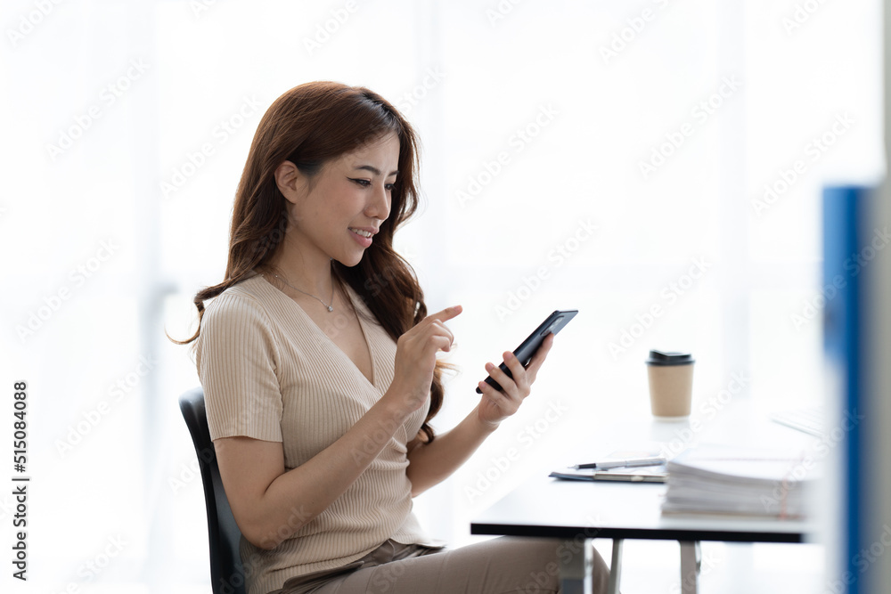 Portrait of smiling young woman with smartphone in her hands, sitting at table in front of laptop. Girl works on computer while sitting in office, checks email. Online marketing, distance education. Portrait of smiling young woman with smartphone in her hands, sitting at table in front of laptop. Girl works on computer while sitting in office, checks email. Online marketing, distance education.