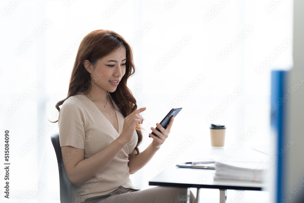 Portrait of smiling young woman with smartphone in her hands, sitting at table in front of laptop. Girl works on computer while sitting in office, checks email. Online marketing, distance education. Portrait of smiling young woman with smartphone in her hands, sitting at table in front of laptop. Girl works on computer while sitting in office, checks email. Online marketing, distance education.