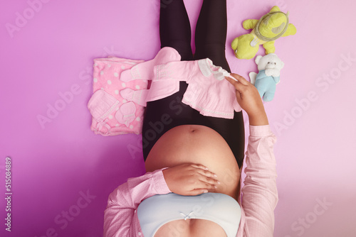 Pregnant woman sitting on floor with baby clothes, in the middle, top view 