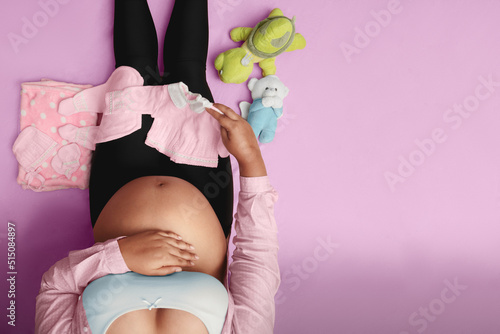 Pregnant woman sitting on floor with baby clothes and stuffed bear, turtle, top view 