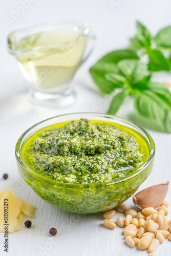 Green fresh homemade italian pesto sauce made of blended basil leaves, olive oil, pine nuts, garlic and parmesan served in transparent glass bowl on white wooden table used in salad or pasta seasoning