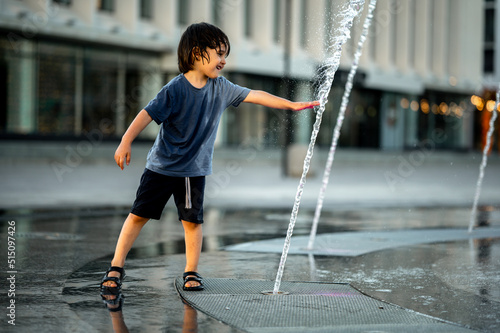 a boy of Asian appearance with long hair in a blue T-shirt is bathing in a fountain on a hot summer day. The kids are having fun . The boy cools down from the heat with the water of the city fountain