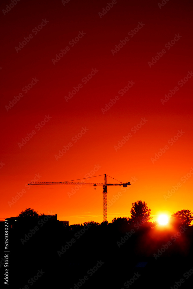 Tall crane silhouette against warm orange sunset sky. City skyline. Sun glow and flare. Dramatic rich saturated color. Tall crane silhouette against warm orange sunset sky. City skyline. Sun glow and flare. Dramatic rich saturated color.