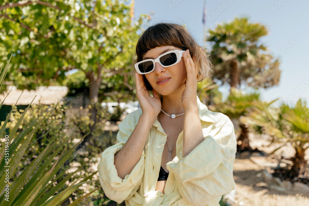 Close up portrait of trendy girl with short hairstyle wetting with sunglasses and summer yellow suit touching er glasses and posing on summer day on background of palm trees Close up portrait of trendy girl with short hairstyle wetting with sunglasses and summer yellow suit touching er glasses and posing on summer day on background of palm trees