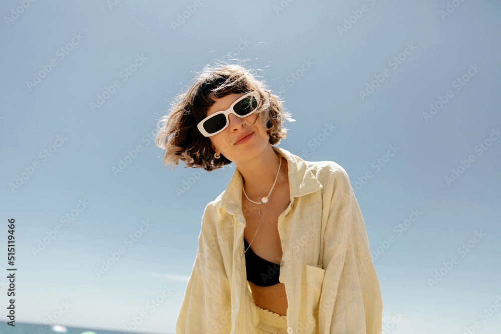 Gorgeous young woman posing emotionally with lovely smile against blue sky in sunny day. Pretty dark-haired girl with short wavy hair in sunglasses and yellow shirt expressing happiness.