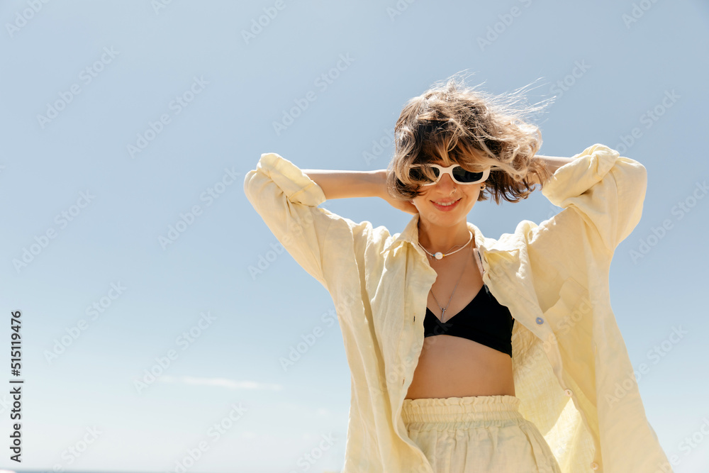 Stylish adorable pretty woman with wavy hair wearing linen yellow suit in sunglasses dancing and having fun against blue sky in summer sunny day