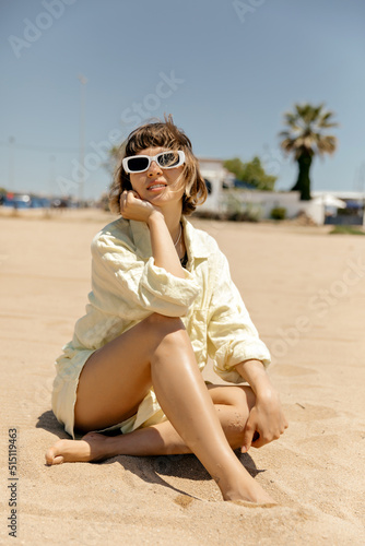 Caucasian young tanned girl is sitting on sandy beach in summer outfit. Blonde with short hair looks back smiling with teeth. Healthy lifestyle people concept.